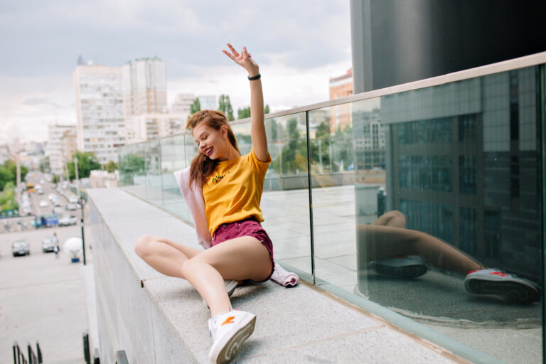 Smiling ginger girl in yellow shirt lying on stone parapet and looking down. Outdoor portrait of young brunette woman in white sneakers having fun alone and enjoys city view in morning.